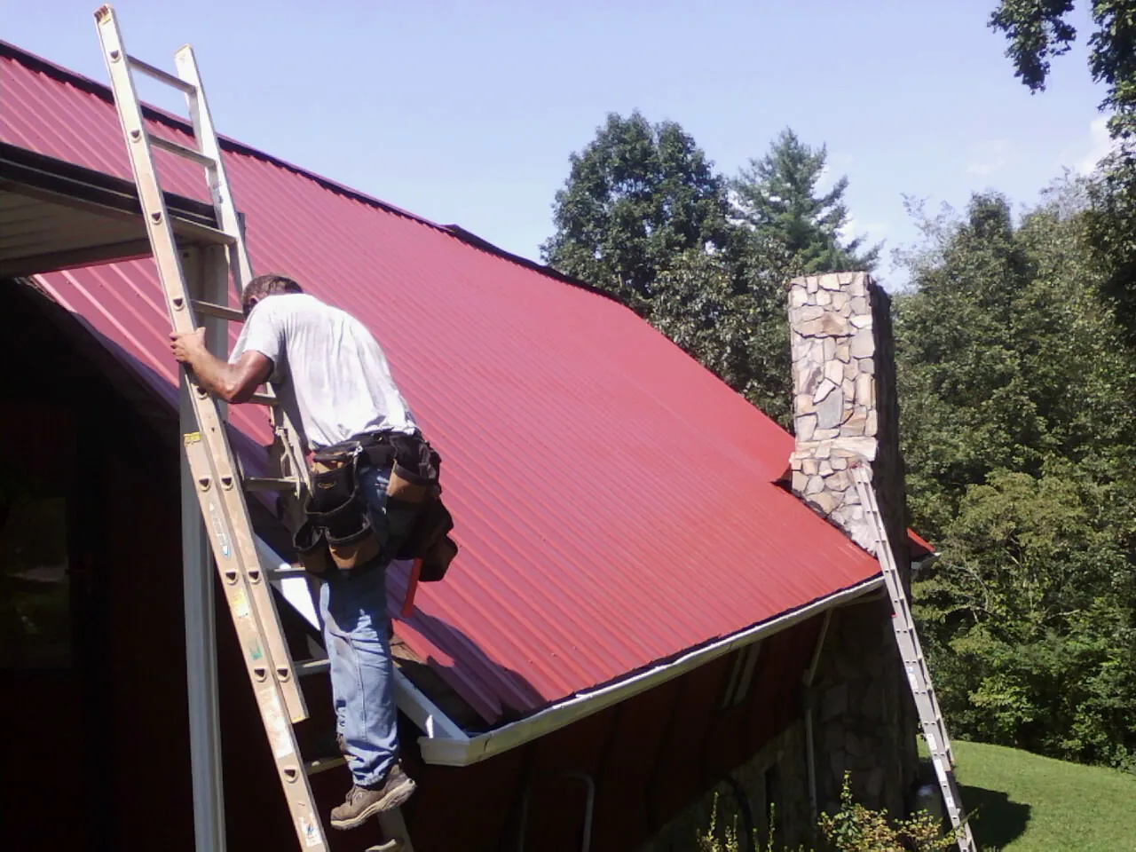 Roofer on a ladder beside a red metal roof and stone chimney — Soffit & Fascia Repair by Roofing Brothers in Oreland